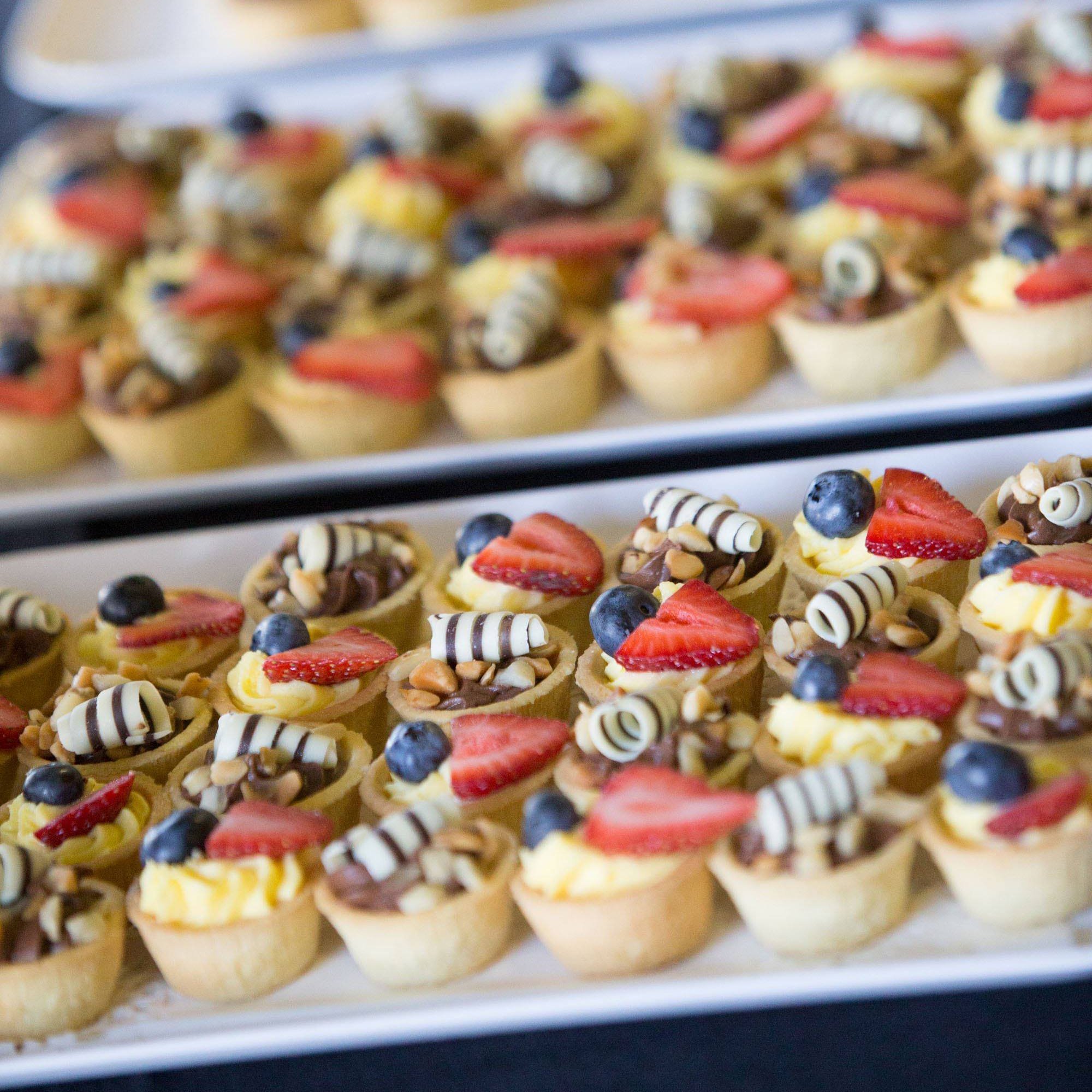 Tray of small fruit tarts. The tarts are arranged in rows on a white tray and have a variety of toppings, including strawberries, blueberries, bananas, and nuts.