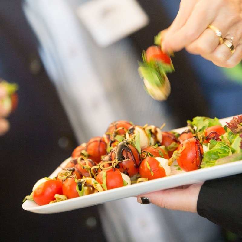 white rectangular plate of appetizers being served by a person. The appetizers are small red tomatoes with green leaves and black sesame seeds on top.
