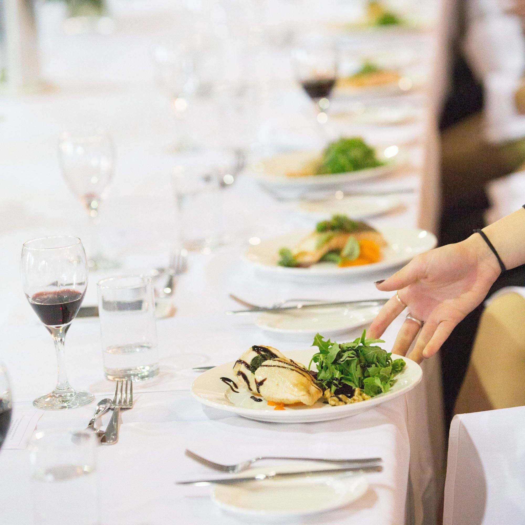 The table is set with a white tablecloth, plates, silverware, and wine glasses. The plate of food appears to be a salad with greens, grilled vegetables, and a protein. The background consists of other tables with similar table settings.